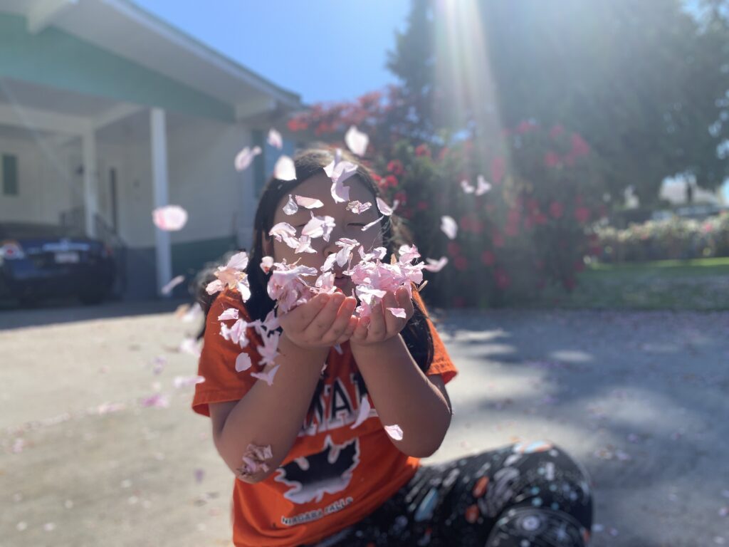 An Asian girl in an orange t-shirt sitting on the pavement in front of a blurred house, blowing cherry blossom petals at the camera