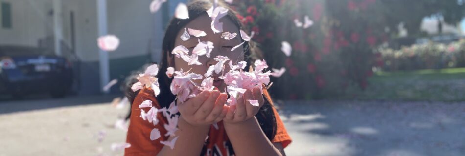 An Asian girl in an orange t-shirt sitting on the pavement in front of a blurred house, blowing cherry blossom petals at the camera