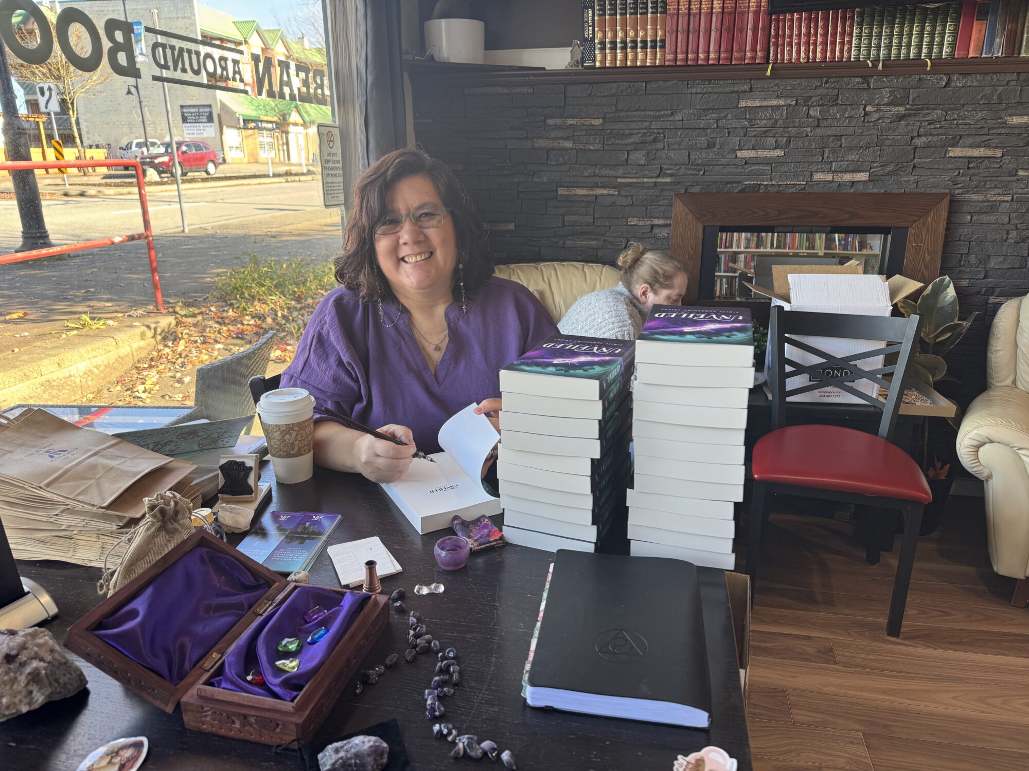 Author, a middle aged woman with wavy dark brown hair and glasses, happily signing a book at a table displaying the book, various amethyst stones, and some stickers and bookmarks