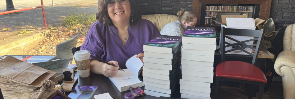 Author, a middle aged woman with wavy dark brown hair and glasses, happily signing a book at a table displaying the book, various amethyst stones, and some stickers and bookmarks